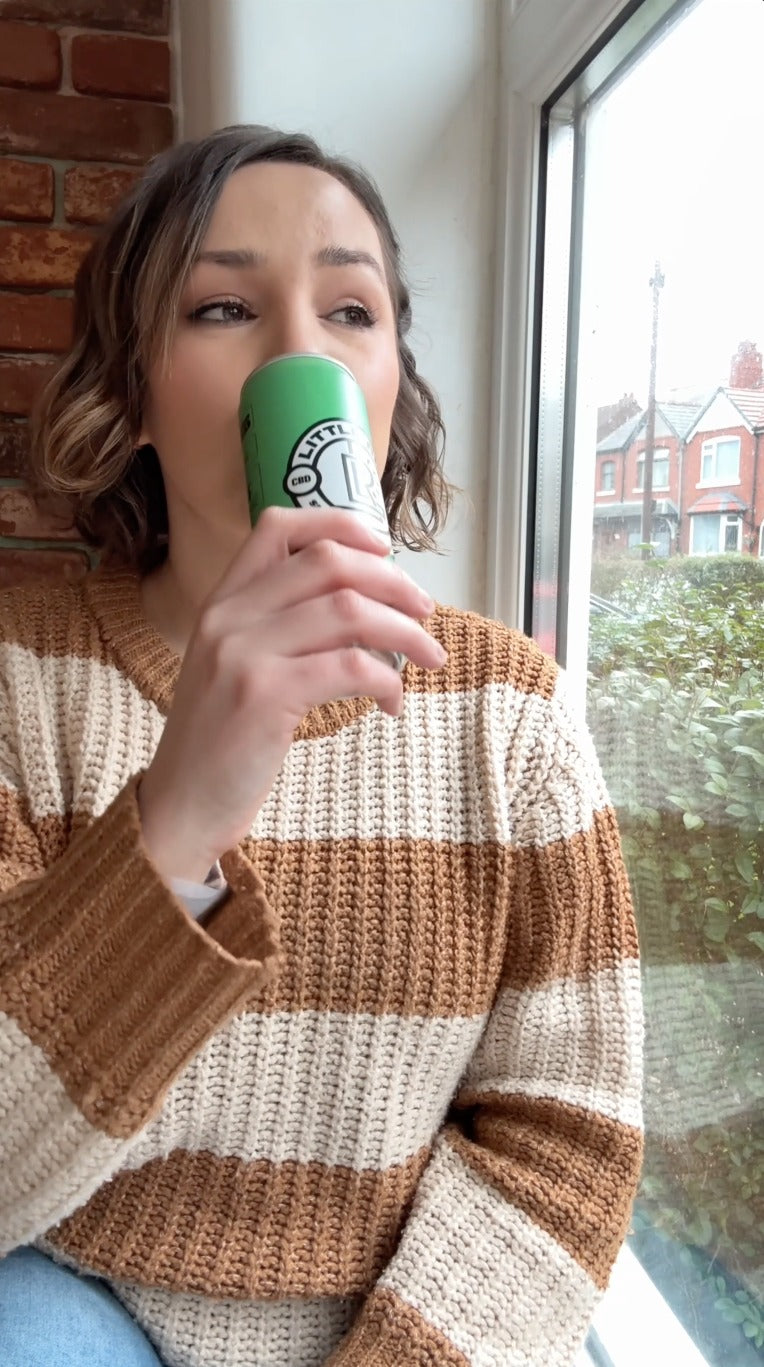 Woman in a striped sweater drinks from a green can while sitting by a window.