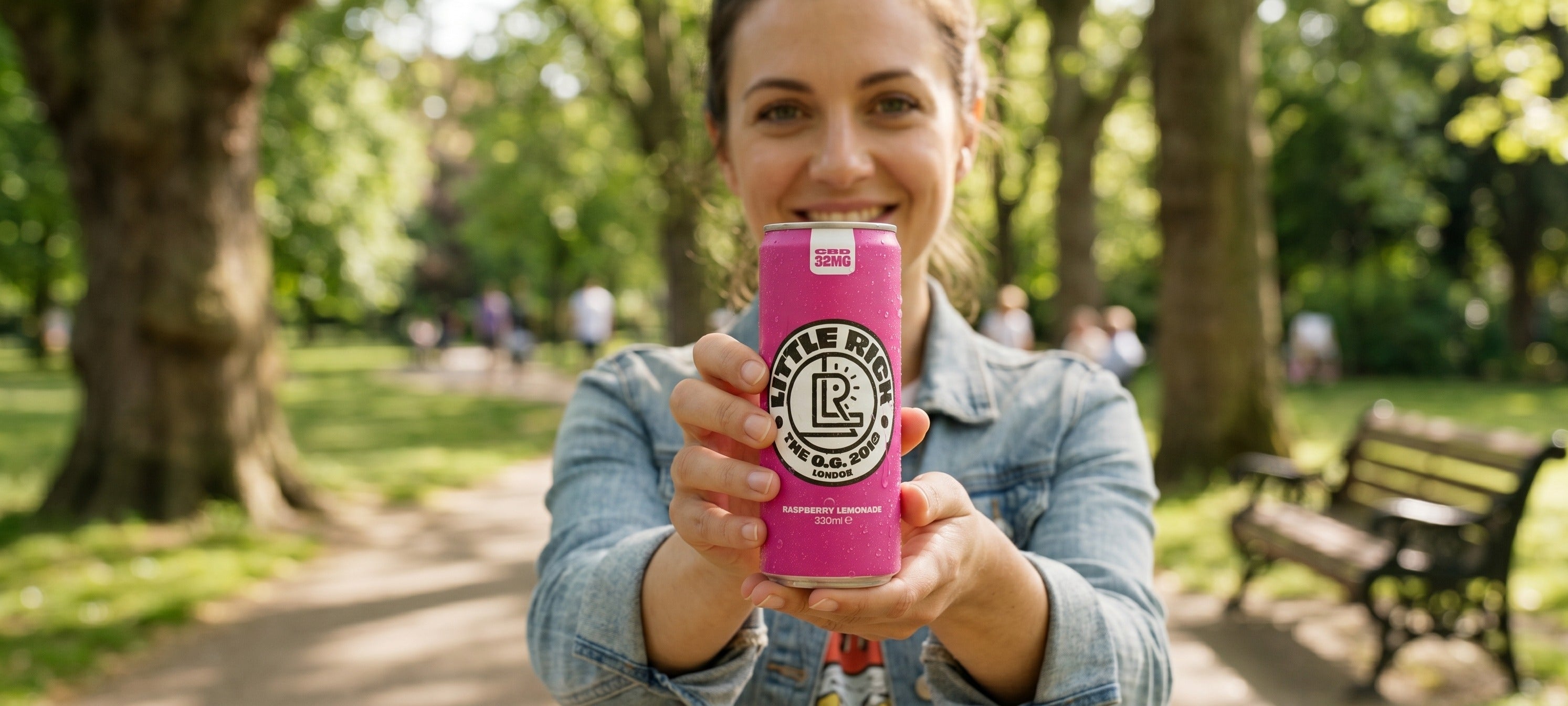 Girl at park with raspberry lemonade can with trees and a bench behind.